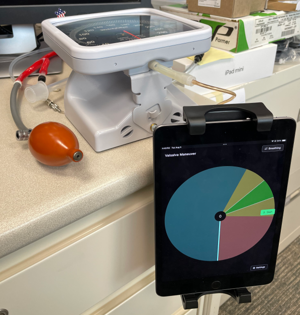 An iPad shows a pressure dial with various colored regions. It is mounted on a desk, which also has a sphygmometer (analog pressure measurement device) and a hand squeeze pump for testing purposes.